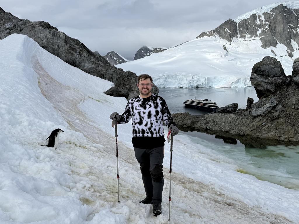 Justin ashore in Antarctica with a penguin and expedition ship behind him