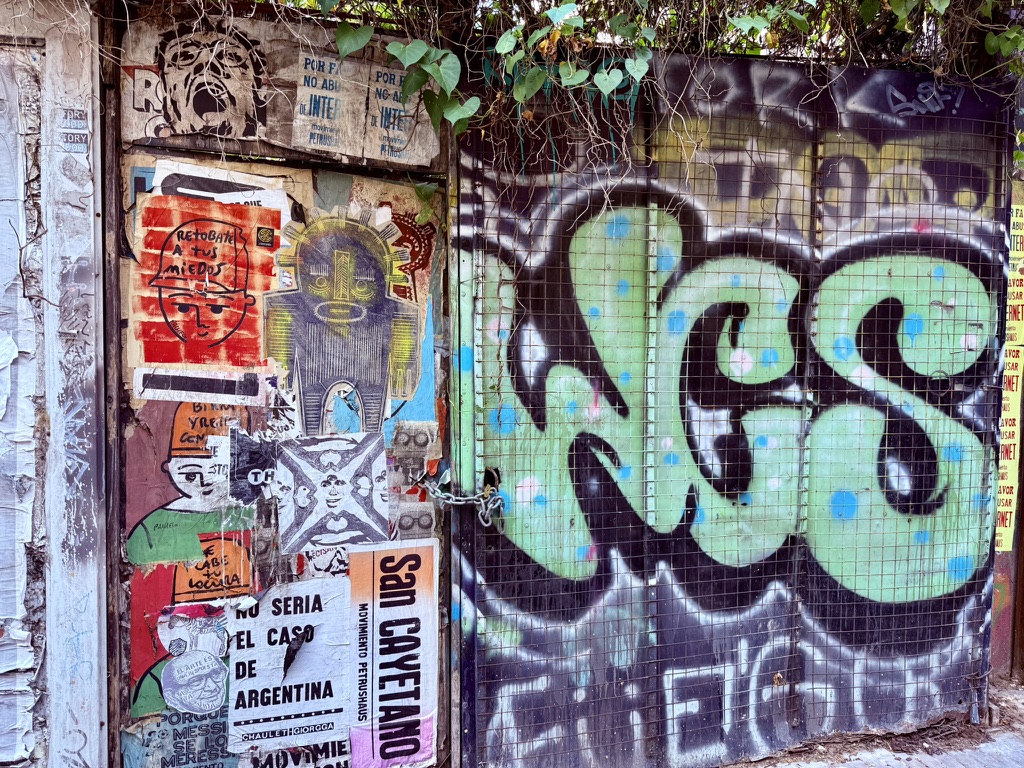A gate covered in green graffiti and colorful posters beneath vines