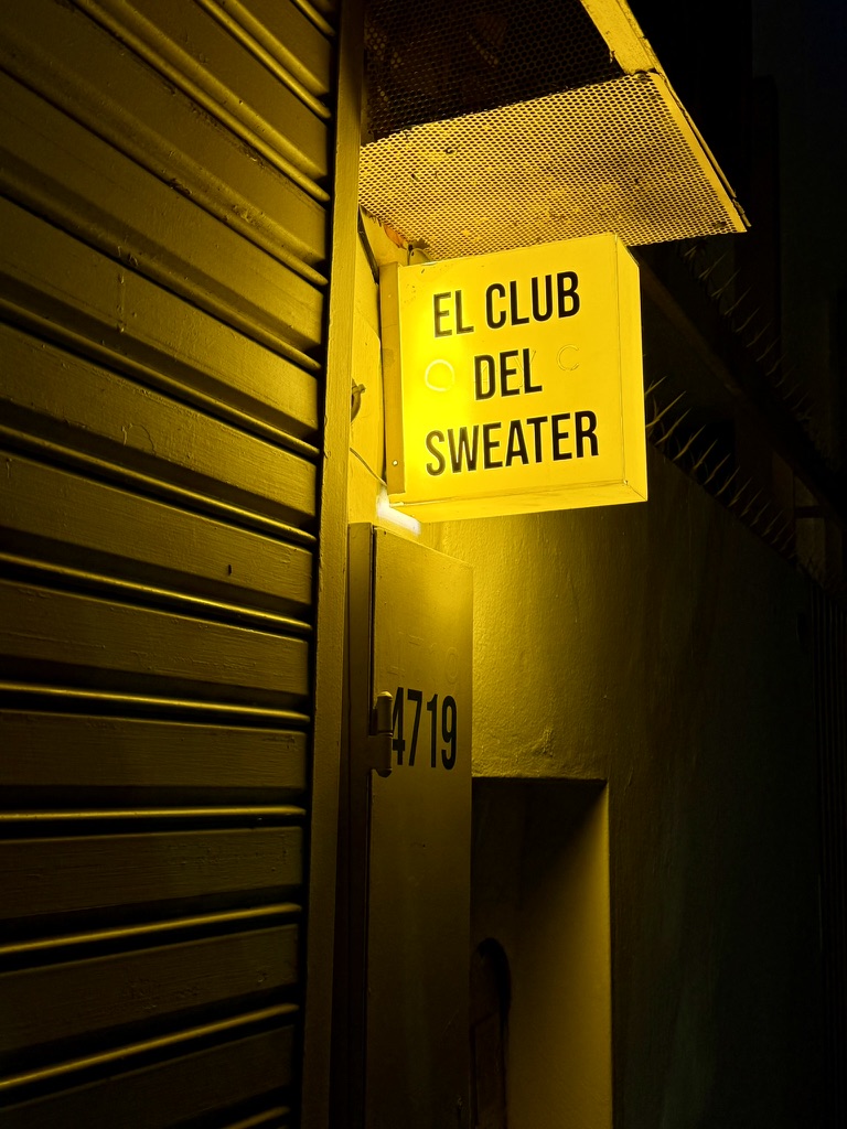 A glowing yellow El Club del Sweater sign on a dark street